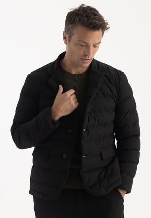 Homme portant une veste matelassée noire par-dessus un pull vert en tricot, regardant vers le bas tout en ajustant le col de la veste d'une main.