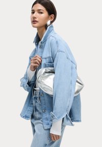 Veste en denim bleu clair avec un large col, associée à un haut blanc ajusté et un jean taille haute, tenant un sac à main en métal argenté.