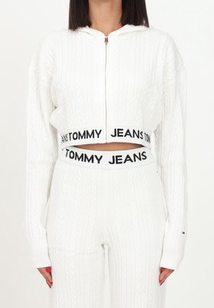 Kobieta w białym, krótkim swetrze na zamek z warkoczowym splotem i dopasowanych spodniach z wysokim stanem z czarnymi taśmami z logo Tommy Jeans.