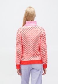 Pletený sveter s jasným korálovým a bielym geometrickým vzorom, s ružovým rebrovaným rolákom a červeným pruhom na leme a manžetách.