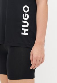 Fekete pamut póló fehér "HUGO" felirattal az oldalán, fekete rövidnadrággal párosítva. Az anyag sima és testhez simuló, alkalmas aktív viseletre.