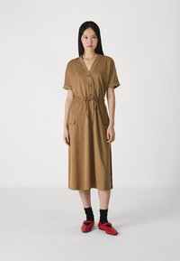 Rochie maro până la genunchi, cu mâneci scurte, cu nasturi pe față și curea la talie; prezintă două buzunare frontale. Asortată cu sosete negre și pantofi roșii.