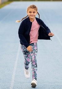 Młoda dziewczynka z warkoczami, ubrana w kwiatowe legginsy i różową koszulę pod czarną kurtką, biegnąca po niebieskim torze.