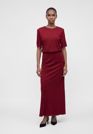 Frau steht, trägt ein langes, eng anliegendes dunkelrotes Kleid mit kurzen Ärmeln und schwarzen High Heels vor einem einfarbigen hellen Hintergrund.