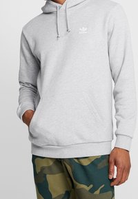 Hombre con sudadera Adidas gris claro con bolsillo frontal y pantalones de camuflaje, con la mano en el bolsillo y en una pose relajada.