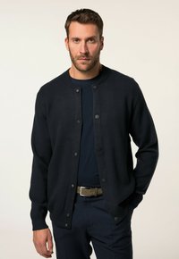 Dunkelmarineblauer Cardigan aus weichem Material, mit rundem Ausschnitt, knöpfbarem Vorderteil und gerippten Bündchen. Kombiniert mit einem marineblauen Hemd und Hose.