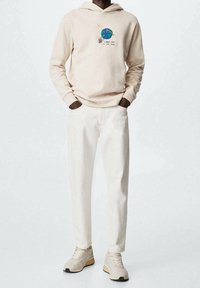 Felpa beige con cappuccio con un disegno grafico di un pianeta e stelle, abbinata a pantaloni bianco sporco e sneakers beige chiaro.