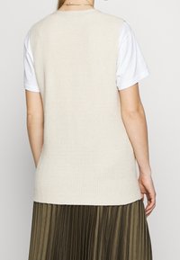 Beige ribbat stickad väst som bärs över en vit t-shirt, tillsammans med en plisserad olivgrön kjol. Vesten har en figursydd silhuett och rund halsringning.