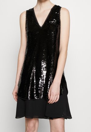 Top nero senza maniche con paillettes e scollo a V indossato sopra una gonna di chiffon nera, che mostra la parte superiore del corpo e le braccia su sfondo semplice.