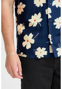 Camicia a maniche corte di colore blu navy con grandi fiori color crema e centri rossi; presenta bottoni neri e un'etichetta laterale; indossata con pantaloni neri.