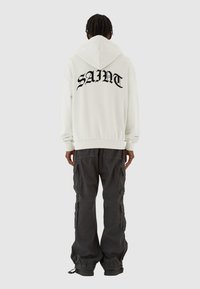 Weißer Kapuzenpullover mit dem Wort "SAINT" in fettem schwarzen Text auf dem Rücken, kombiniert mit dunklen Cargo-Hosen mit Taschen und verstellbaren Bündchen.