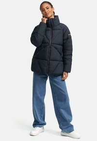 Puffjakke i mørkeblå, med høj krave, quiltet mønster og frontlynlås. Sammen med løse blå jeans og hvide sneakers.