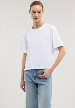 Mujer vistiendo una camiseta blanca de manga corta lisa y jeans de mezclilla azul claro, de pie con una mano en el bolsillo.