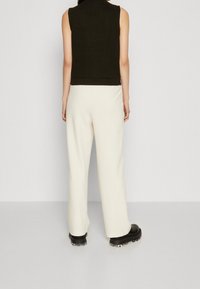 Schwarzes ärmelloses Oberteil mit strukturierter Oberfläche, kombiniert mit cremefarbenen Hosen mit weitem Bein und klobigen schwarzen Schuhen mit transparentem Sohlenbereich.