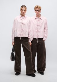Δύο άτομα φορούν πανομοιότυπα ροζ ανοιχτά πουκάμισα με υφή και διακοσμητικές γραβάτες, σε συνδυασμό με καφέ σκούρα παντελόνια φαρδιάς γραμμής και μαύρα παπούτσια.
