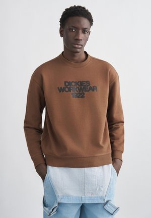 Mladý muž oblečený v hnedom pulóvri s nápisom "Dickies Workwear 1922" a svetlomodrých džínsových náprsenkových nohaviciach s rukami v vreckách, neutrálne pozadie.