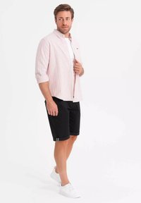 Chemise à rayures roses et blanches, portée ouverte sur un t-shirt blanc, assortie à un short noir et des baskets blanches. Design simple et décontracté.
