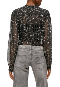 Kvinde iført en gennemsigtig sort blomsterbluse med lange puffede ærmer, stoppet ind i højtaljede slidte grå jeans, set fra ryggen.