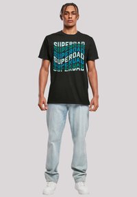 Black t-shirt featuring "SUPERDAD" text in turquoise and light blue, with a bold design. Paired with light blue jeans and white sneakers.