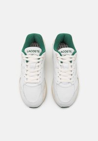 Białe sneakersy z zielonymi akcentami, posiadające teksturowaną siateczkową cholewkę, okrągły czubek i sznurowane zapięcie. Nazwa marki "LACOSTE" na języku.
