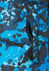 Přechodná bunda v modrém maskáčovém vzoru s černými doplňky. Má kapsu na zip a hladkou, vycpanou strukturu. Odolná tkanina.