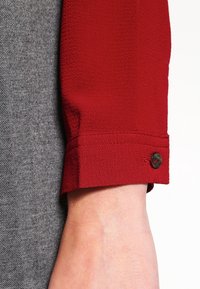 Manche en tissu rouge à finition texturée, avec un détail de bouton et un poignet effilé, adjacente à un vêtement gris texturé.