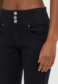 Črne hlače iz jeansa z visokim pasom, tremi srebrnimi gumbi in sprednjimi žepi. Tkanina je gladka na otip, dizajn je oprijet.