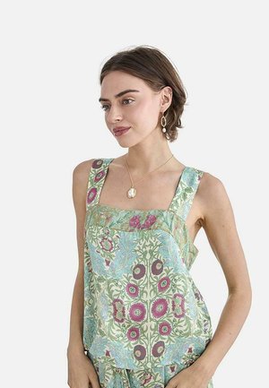 Mujer con cabello corto y castaño que lleva un vestido sin mangas con estampado floral en verde y púrpura, collar con colgante dorado y pendientes colgantes.