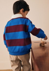 Modro-vínová pruhovaná mikina s bílým límečkem, doplněná béžovými kalhotami. Dítě má zády otočeno a je zapojené do hry s dřevěným modelovým letadlem.