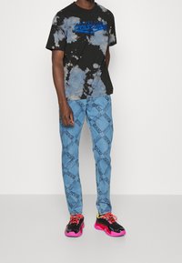 Schwarzes Batik-T-Shirt mit blauem Logo, kombiniert mit hellblauen Hosen, die ein wiederholendes Textmuster aufweisen. Schwarze Sneaker mit pinkfarbenen Sohlen.
