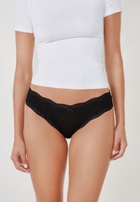 Femme portant un bikini noir orné de dentelle et un haut blanc moulant à manches courtes, debout devant un fond uni.