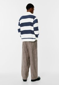 Prugasti pulover u mornarskoj plavoj i bijeloj, od pamučne mješavine, s opuštenim krojem. Uparen s hlačama širokih nogavica od teksturiranog smeđeg materijala i crnim cipelama.