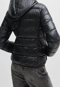 Sort, skinnende pufferjakke med horisontalt quiltet mønster, hætte og elastiske manchetter, parret med grå, højtaljede jeans.