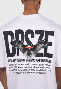 Hvit bomullst-skjorte med et stort svart ørnegrafikk og fet tekst som sier "DPSZE" og "REALITY IS WRONG, DREAMS ARE FOR REAL."