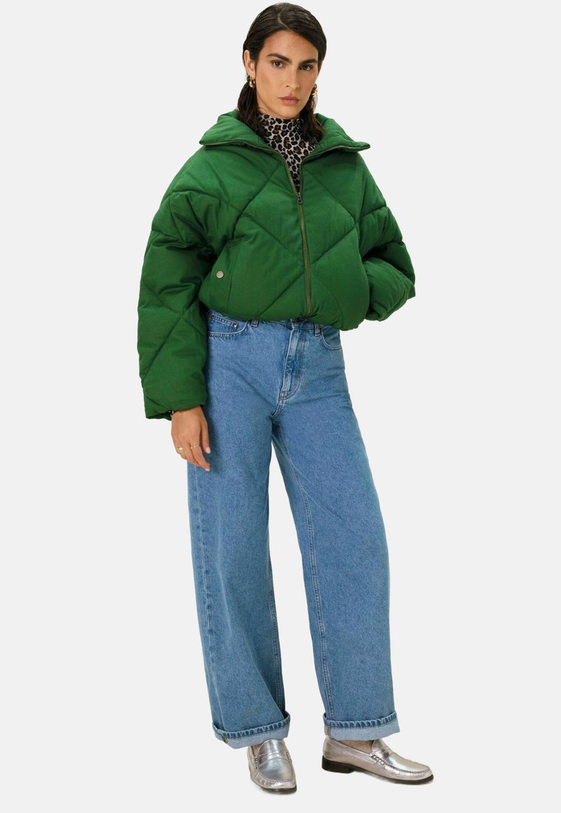 Veste matelassée verte avec fermeture éclair, associée à un jean large taille haute bleu et des mocassins argentés brillants. Comprend un col roulé à motifs.