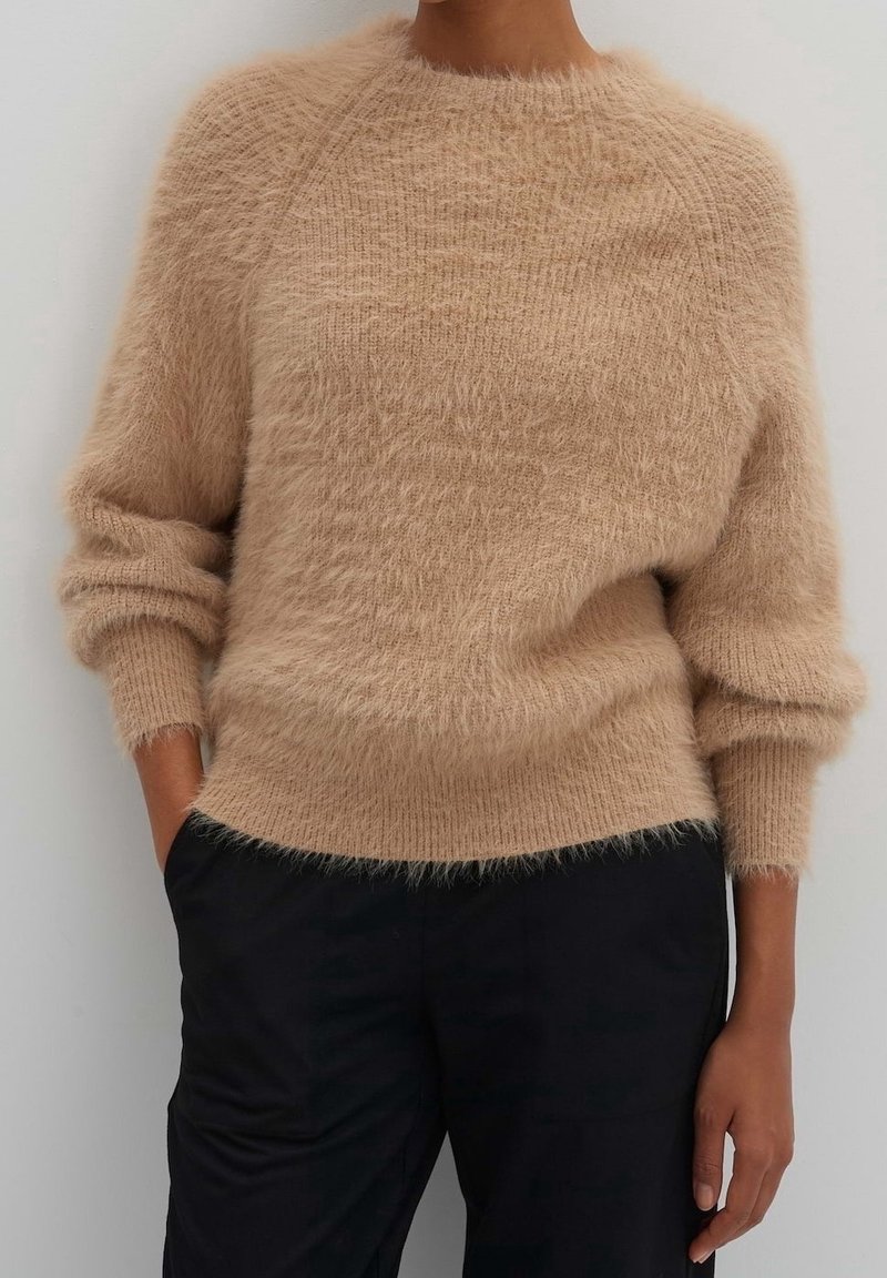 Personne portant un pull en maille beige duveteux à manches longues et un pantalon noir, debout avec une main dans la poche sur un fond uni.