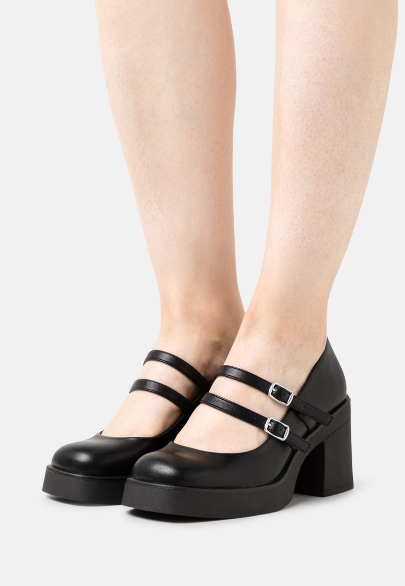 Paire d'escarpins Mary Jane en cuir noir à plateforme avec double brides et boucles argentées, portés sur pieds nus.