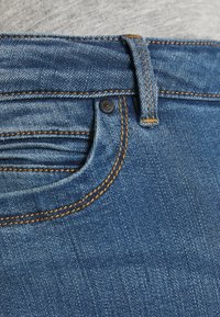 Los vaqueros de mezclilla azul oscuro cuentan con un cierre de cremallera, diseño estándar de cinco bolsillos y costuras en naranja contrastante en las costuras.