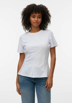 Kvinde med krøllet hår iført en tætsiddende hvid top med korte ærmer og blå jeans, stående mod en ensfarvet hvid baggrund.