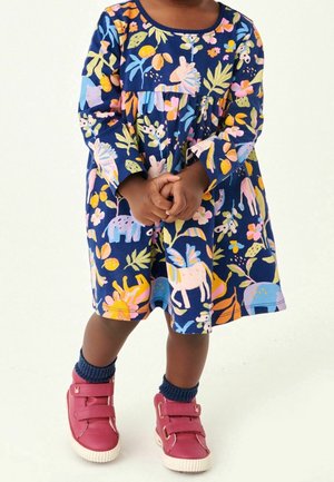 Vestido azul marino con mangas largas, que presenta patrones coloridos de animales y flores. Combinado con zapatos rosas de velcro y calcetines azul marino.