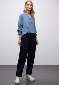 Blusa azzurro chiaro a maniche lunghe con scollo a V e polsini arricciati, abbinata a pantaloni sartoriali blu navy e sneakers bianche.