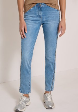 Personne portant un jean droit bleu clair avec un haut marron et des baskets argentées, debout sur un sol clair devant un fond uni.