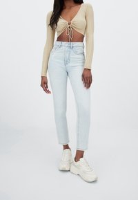 Top corto de canalé beige con lazo en la parte delantera, combinado con jeans de tiro alto azul claro y zapatillas deportivas blancas de suela gruesa. Fondo plano.