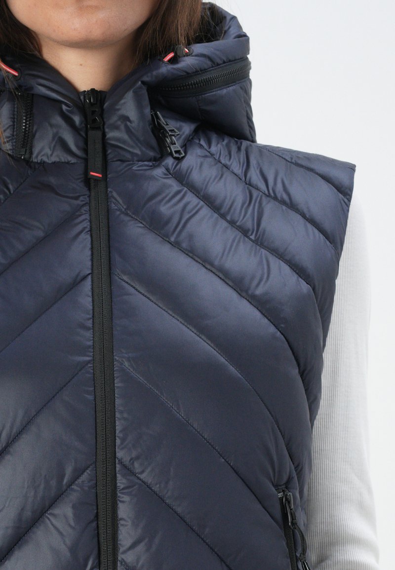 Navyblaue Steppweste mit einem Chevron gesteppte Muster, hohem Kragen und Reißverschluss-Taschen. Verfügt über einen schwarzen Frontreißverschluss und rote Akzente.