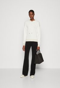 Felpa bianca con cappuccio e finitura testurizzata, abbinata a pantaloni neri a gamba larga e una piccola borsa nera. Sneakers in un colore chiaro completano il look.