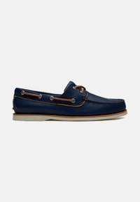 Chaussure bateau en cuir bleu marine avec des lacets couleur tan et une semelle blanche, présentée sur un fond gris clair uni.