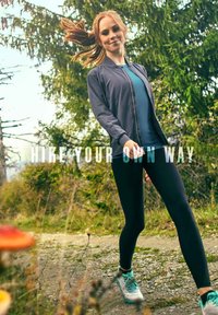 Gray zip-up jacket over a blue shirt, black leggings, and light blue athletic shoes. The background features greenery and a walking path.
