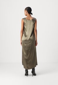 Robe en satin vert olive, longue, avec un décolleté arrondi et des fentes sur les côtés, associée à des bottes à talons hauts noires. Texture lisse et design sans manches.
