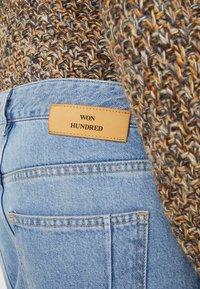 Jeans de mezclilla azul deslavados con una etiqueta de cuero color beige que dice "WON HUNDRED". Suéter marrón de punto detallado parcialmente visible en el fondo.