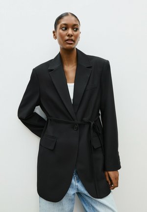 Black blazer with a tie belt, featuring a single button, two front pockets, and a tailored fit, paired with light blue denim.
